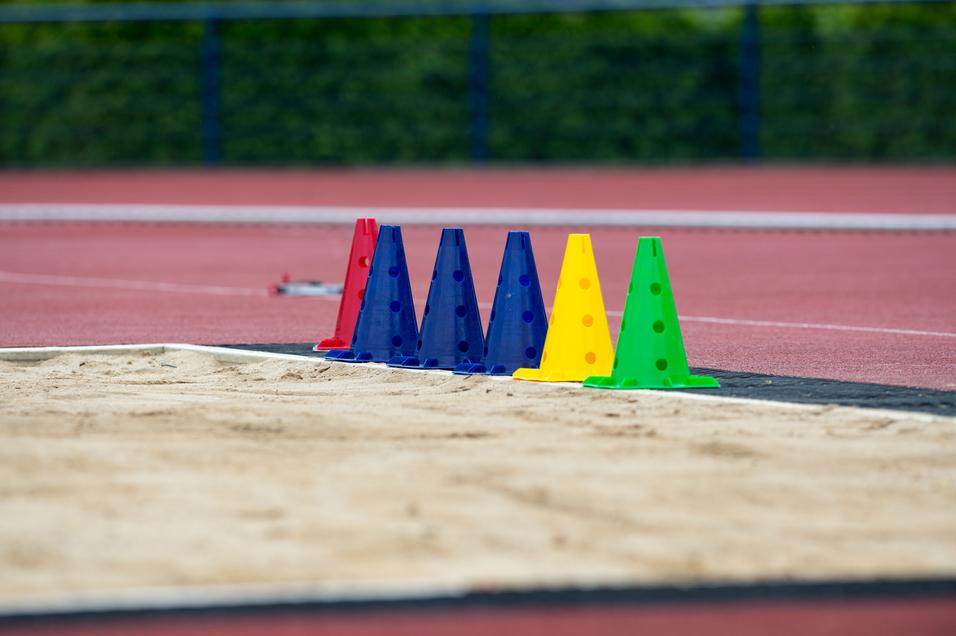 Bunte Trainingshütchen in Rot, Blau, Gelb und Grün stehen neben einem Sandbereich auf einer Sportanlage.