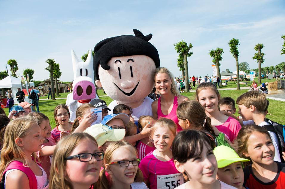 Gruppe von Kindern in pinken T-Shirts posiert lächelnd mit einem Maskottchen und einer Frau auf einer Sportveranstaltung im Freien.